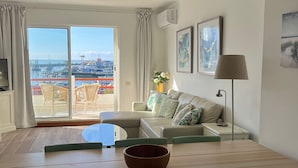 Ein modernes Wohnzimmer mit Essbereich, großen Fenstern und Blick auf eine Marina, einer Couch mit Kissen, einem Couchtisch und einem Fernseher.