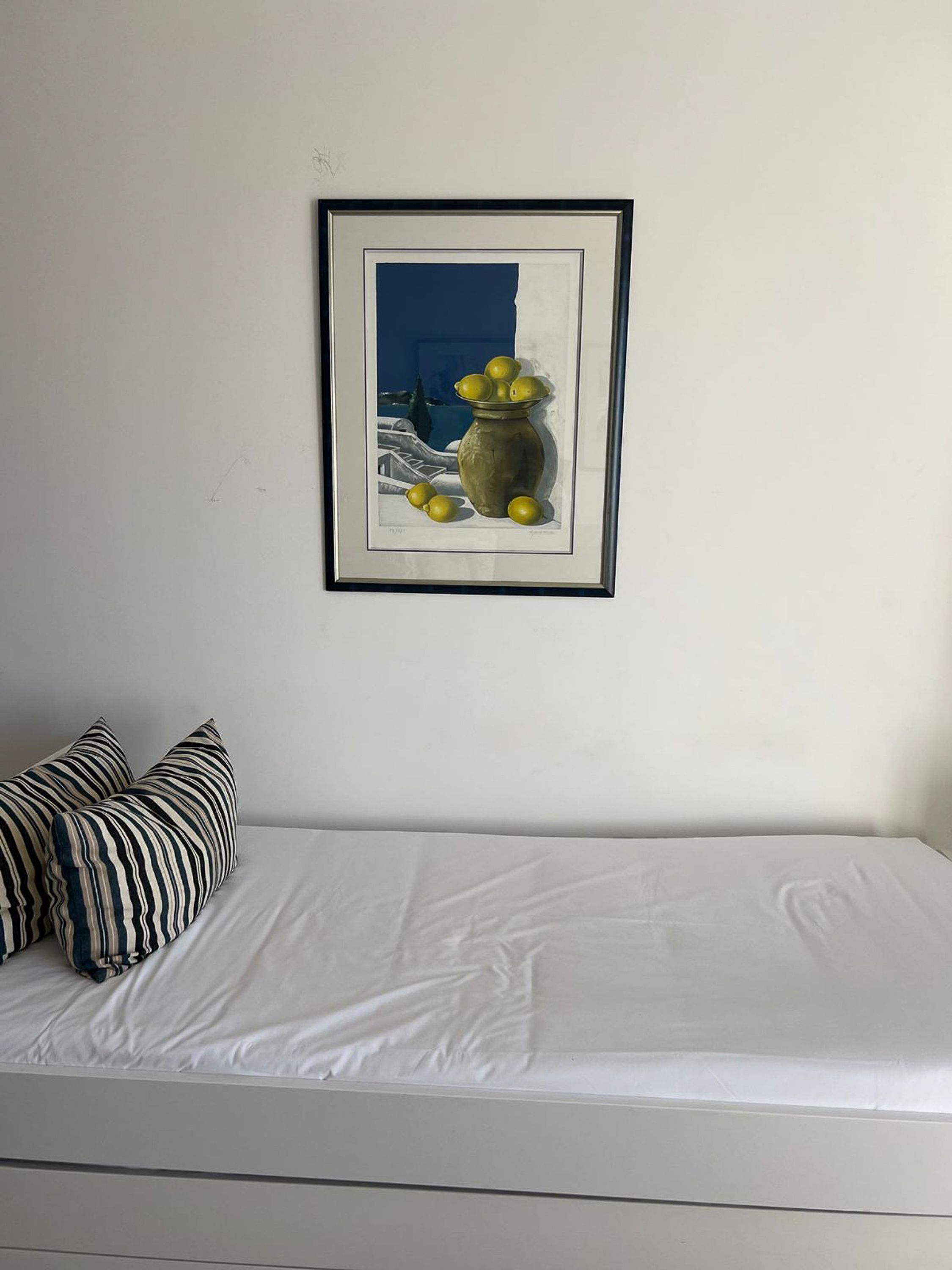Une chambre à coucher avec un lit, un tableau encadré représentant des citrons et un vase, ainsi que des coussins à rayures.