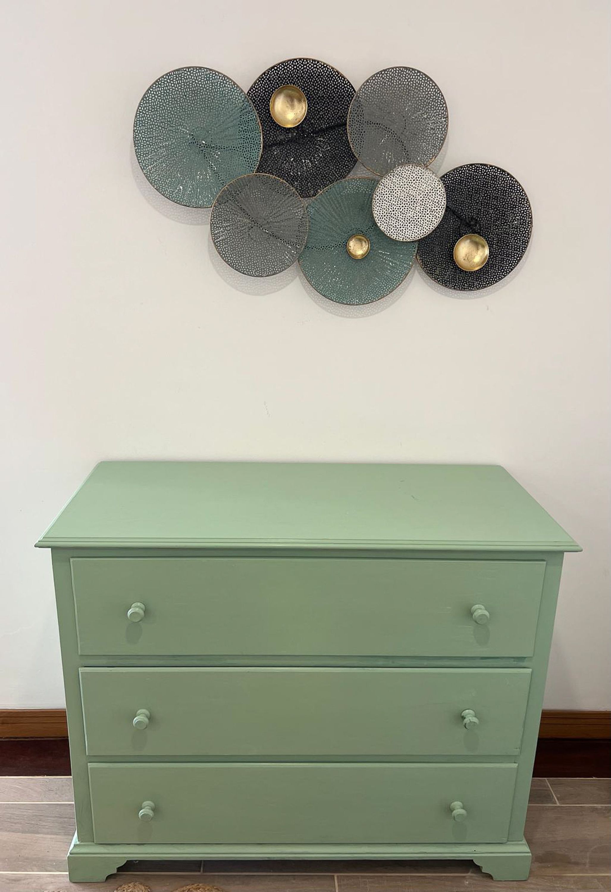 A green dresser with three drawers and a wall-mounted decorative plate arrangement.