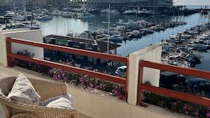 Ein Balkon mit Korbmöbeln, der auf einen Hafen mit Booten und einem Hotel im Hintergrund blickt.