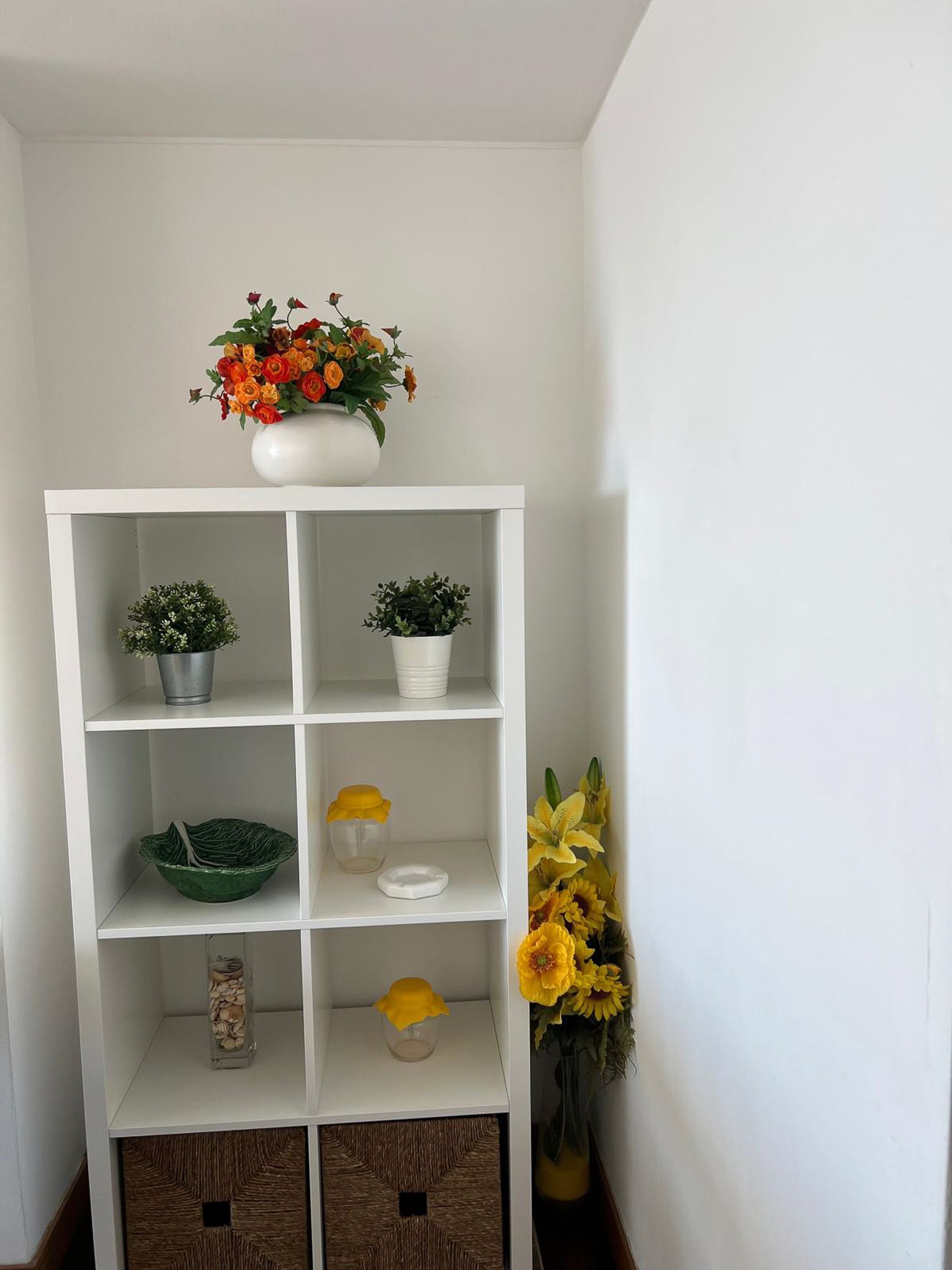 Una estantería blanca con diversos objetos decorativos, entre ellos flores y pequeñas macetas.