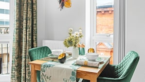 Un comedor con una mesa de madera, sillas tapizadas en verde, un mantel blanco y un florero con flores.