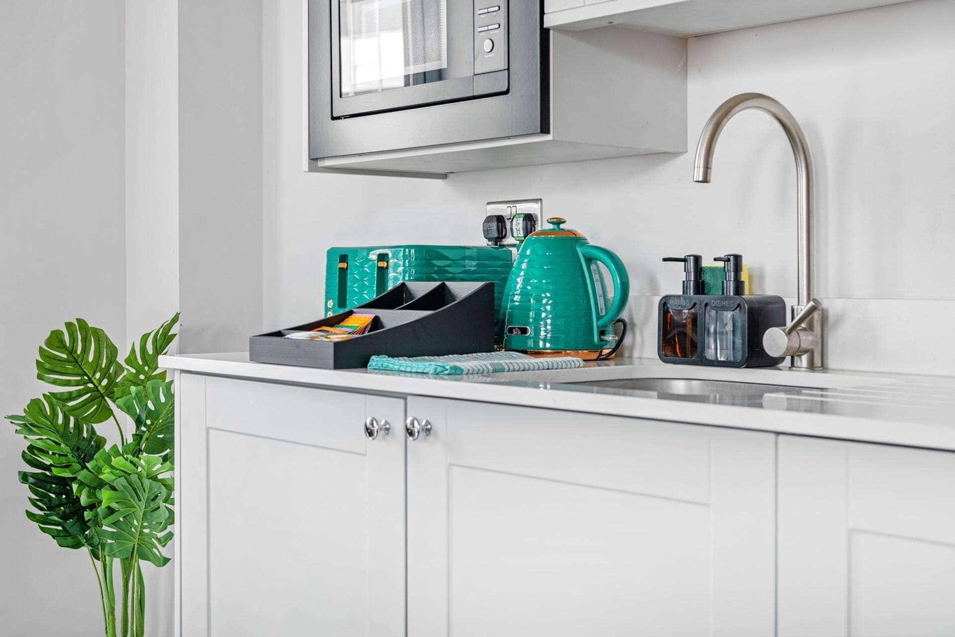 A kitchen with white cabinets, a stainless steel sink, a green kettle, and a microwave above the counter.