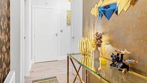 Un pasillo con una puerta blanca, una mesa de vidrio con adornos y una pared texturizada con formas geométricas doradas.