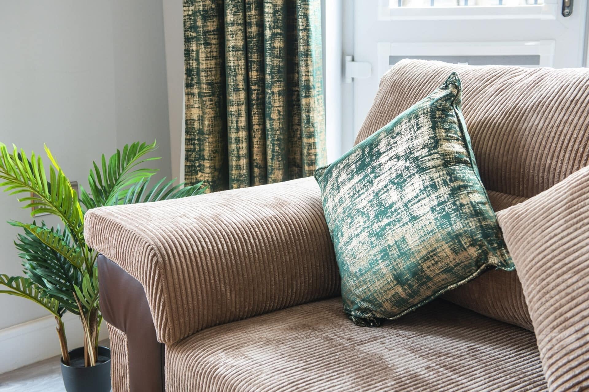 Un sofá color beige con textura y una almohada verde y beige. Se ve una planta en maceta a la izquierda.