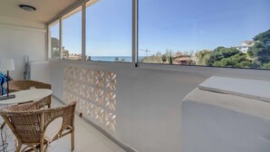 Uma varanda com mobiliário de vime, uma mesa branca e vista para o mar e edifícios.