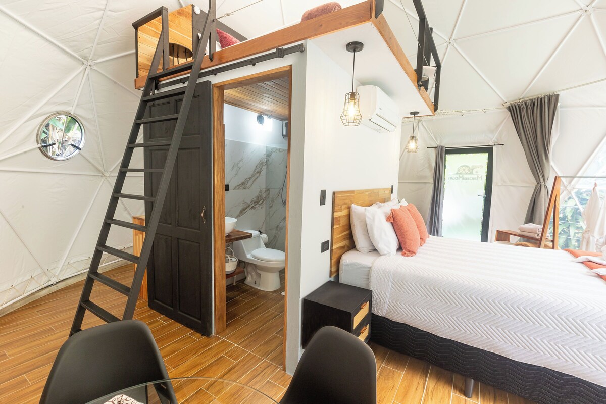 A compact, modern bedroom with a lofted sleeping area, a wooden ladder, a small bathroom, and a large bed with a herringbone-patterned duvet.