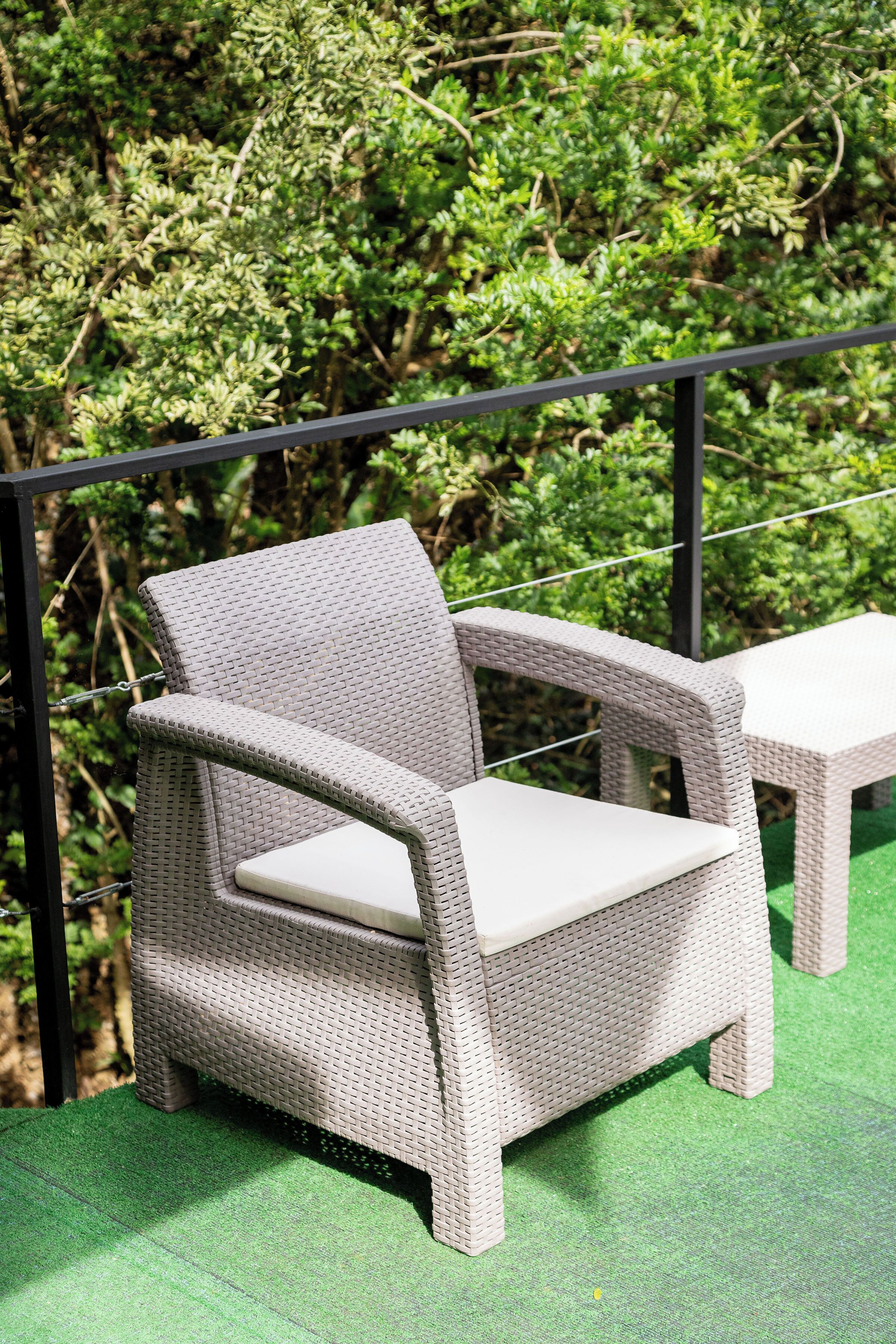 Una silla de mimbre con cojín, sobre una superficie verde, probablemente un patio o terraza, con una barandilla de metal al fondo.