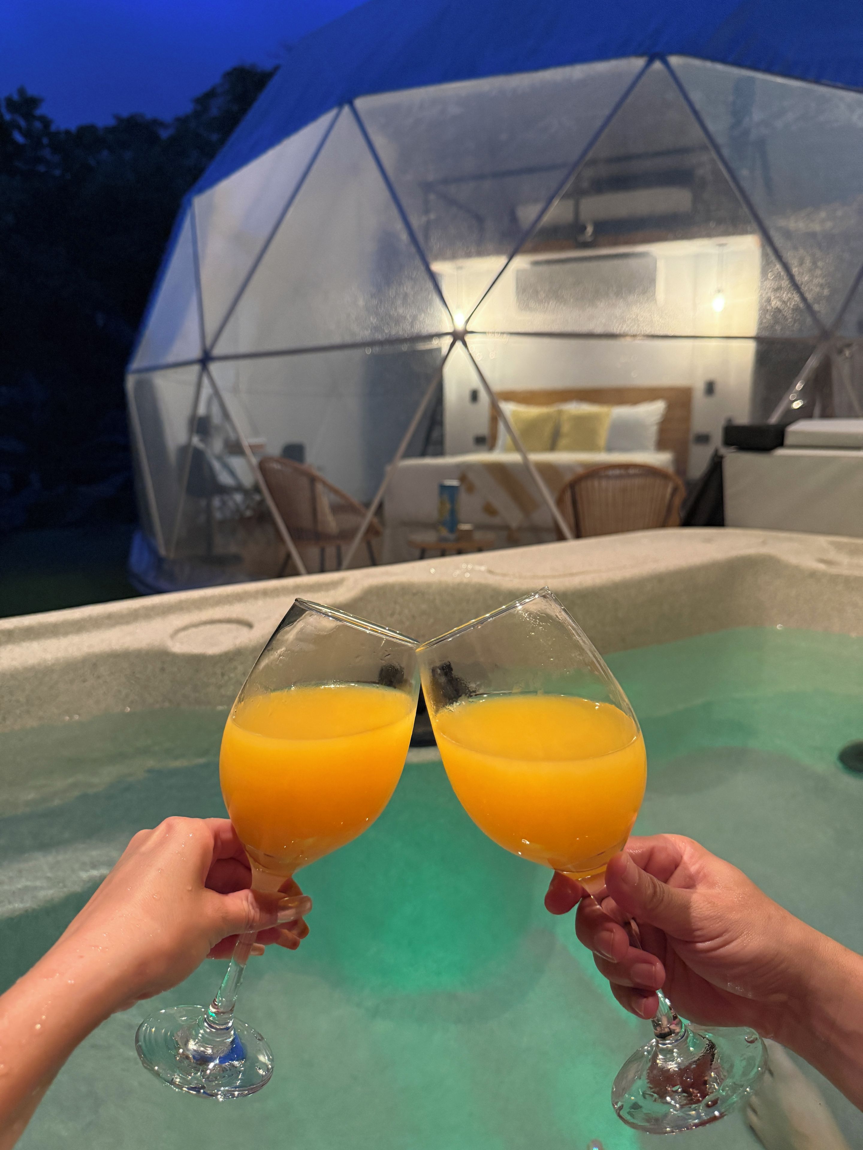 Two hands holding glasses of orange juice in front of a transparent dome structure with a pool.