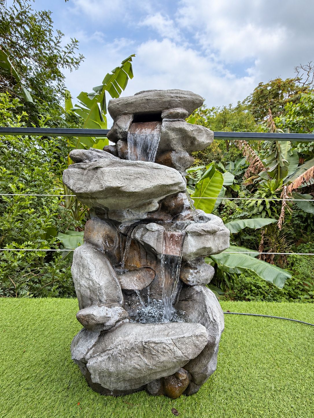 Une cascade artificielle en roche intégrée à un jardin luxuriant, avec une végétation abondante et un ciel dégagé.