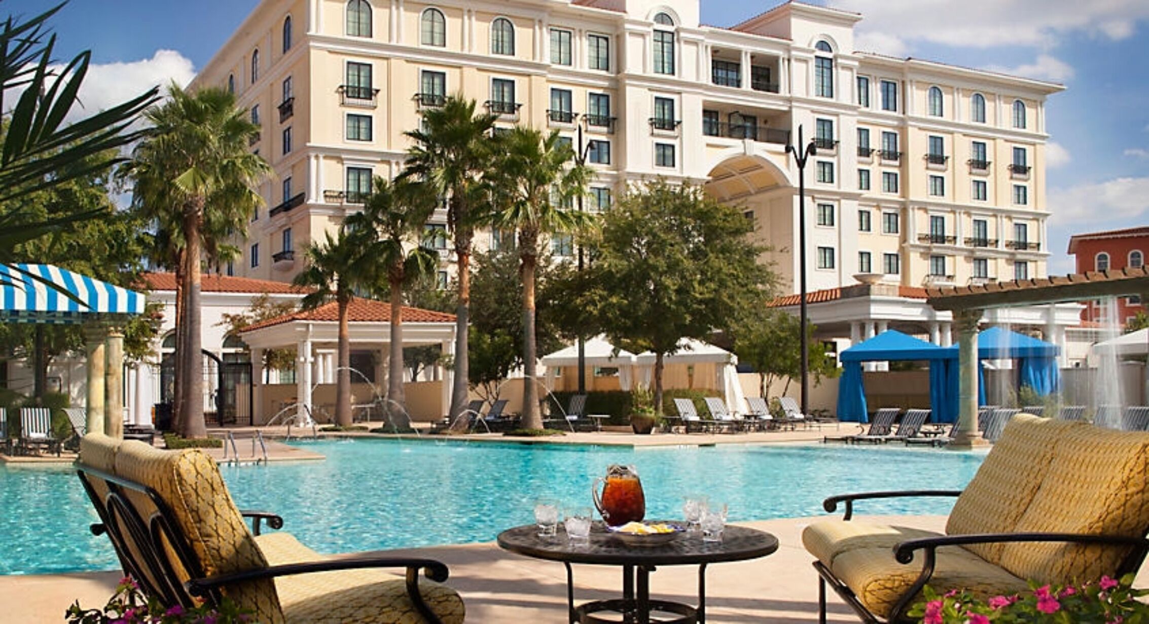 Zona de piscina hotelera con sillones de descanso, una mesa con bebidas y palmeras.