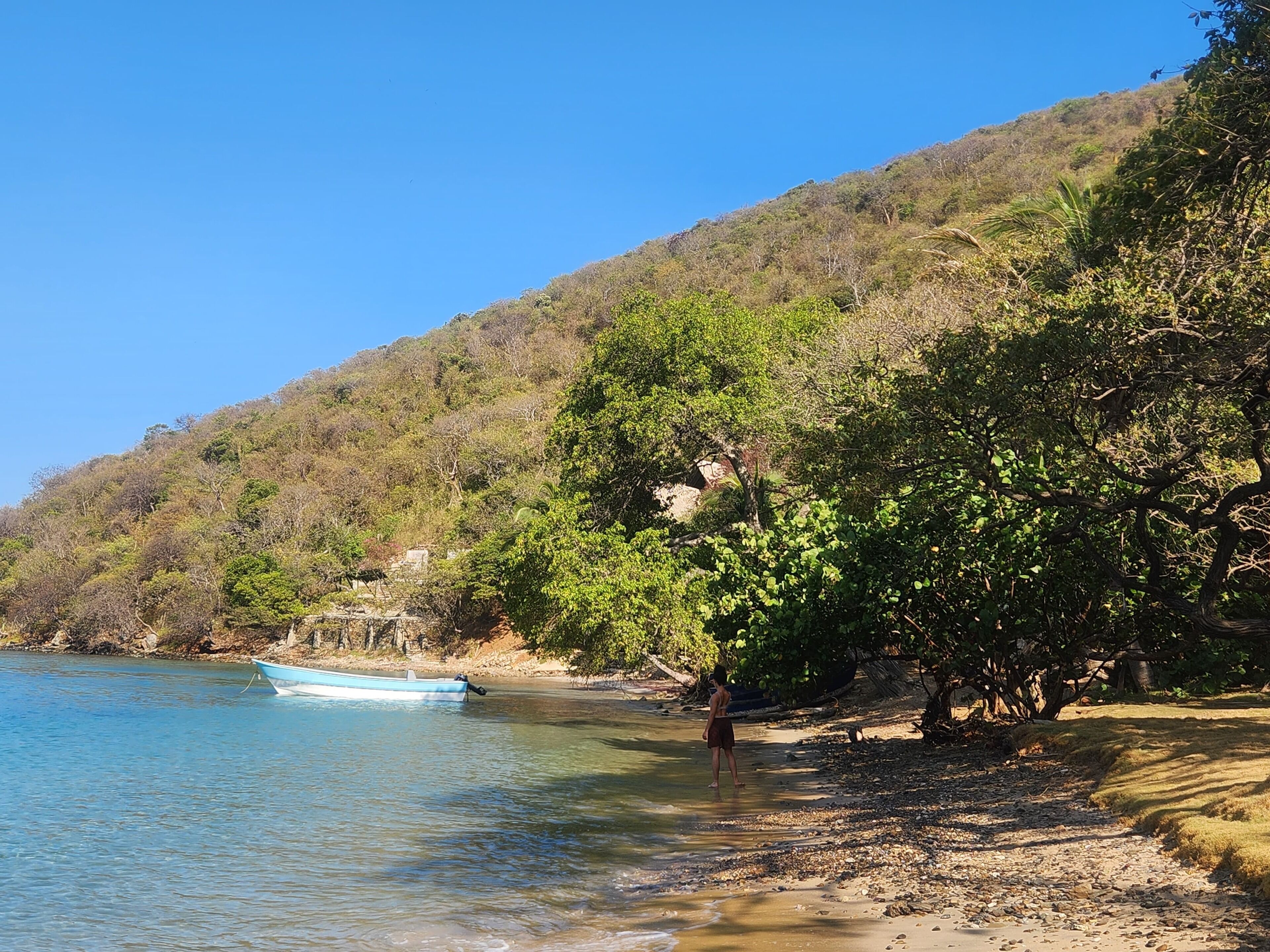 A clear view of a riverbank with a boat docked on the left, a person standing near the water, and a hillside with dense vegetation.
