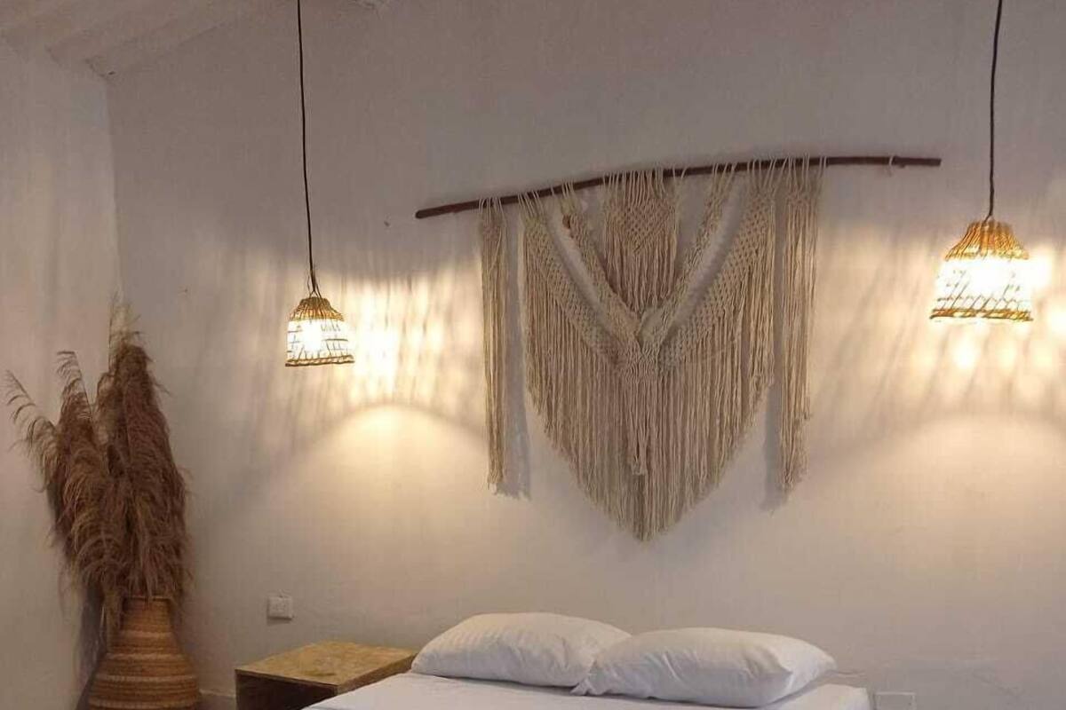 Un dormitorio con cama de madera, ropa de cama blanca, un tapiz de macramé y dos lámparas colgantes.