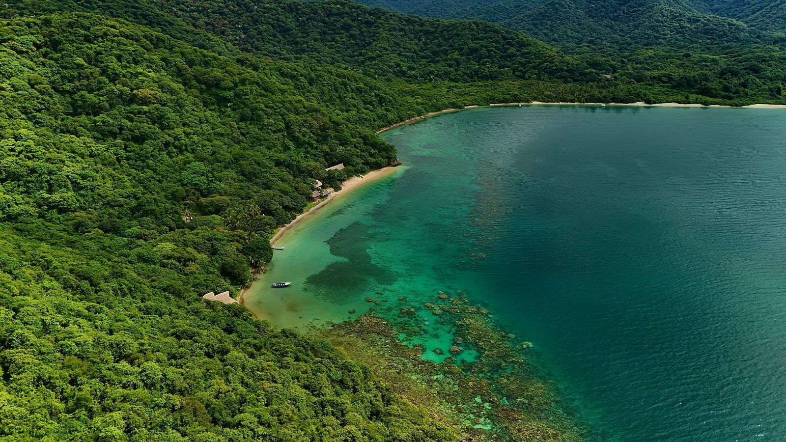 An aerial view of a coastal area with dense green forest, a sandy beach, and clear turquoise waters.