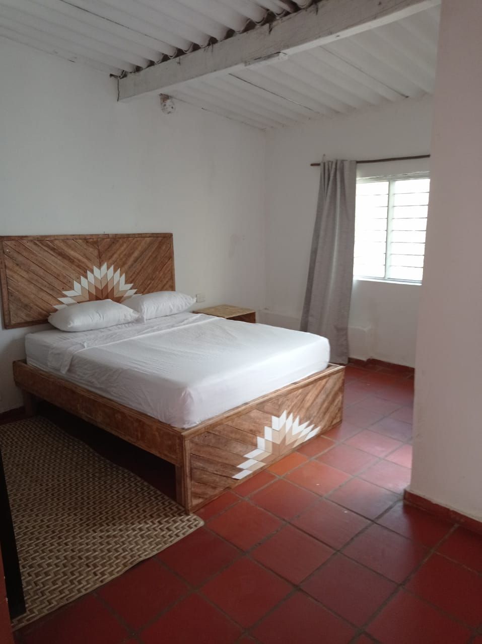 A bedroom with a wooden bed frame, white bedding, a woven rug, and a window with curtains.
