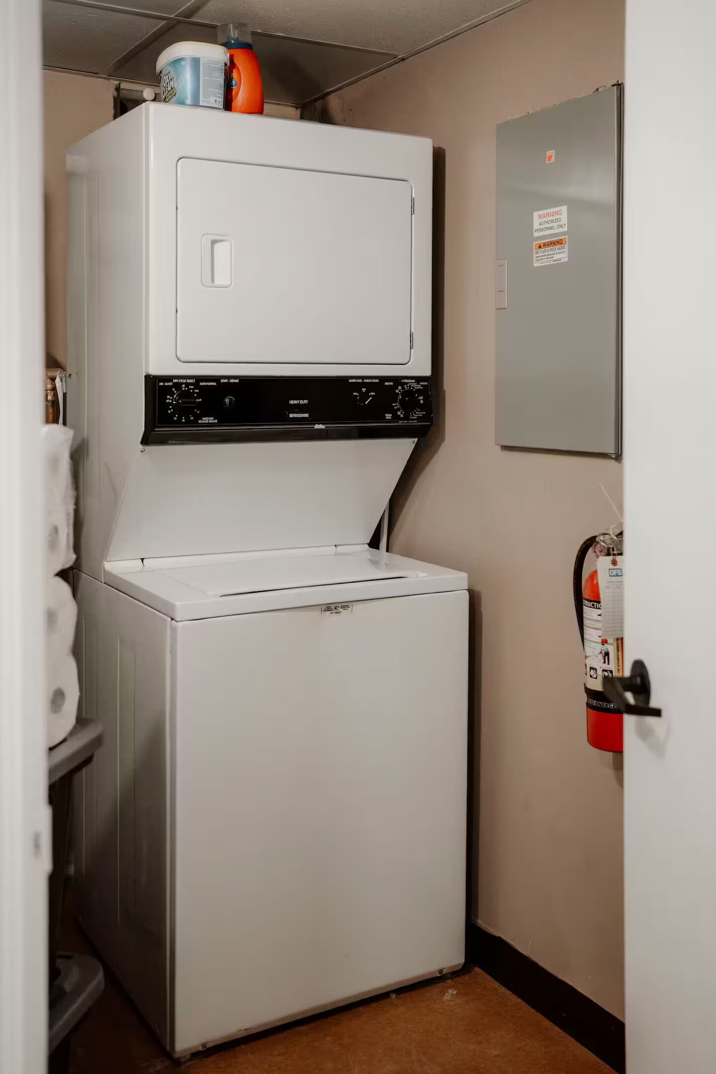 A compact laundry room with stacked appliances, a fire extinguisher, and a warning sign on the wall.