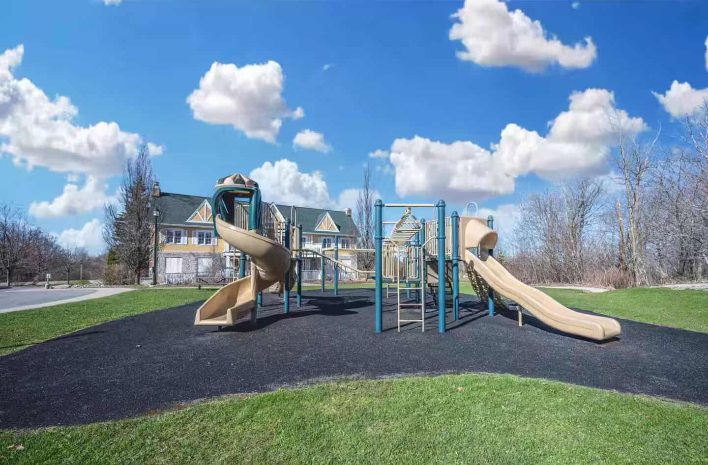 Un parque infantil con dos toboganes y estructuras para escalar, con una casa y un cielo despejado de fondo.