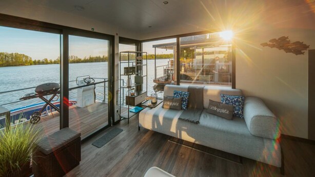 Un salon moderne avec une grande fenêtre donnant sur un plan d’eau, un canapé agrémenté de coussins décoratifs, une petite table et un barbecue à l’extérieur.