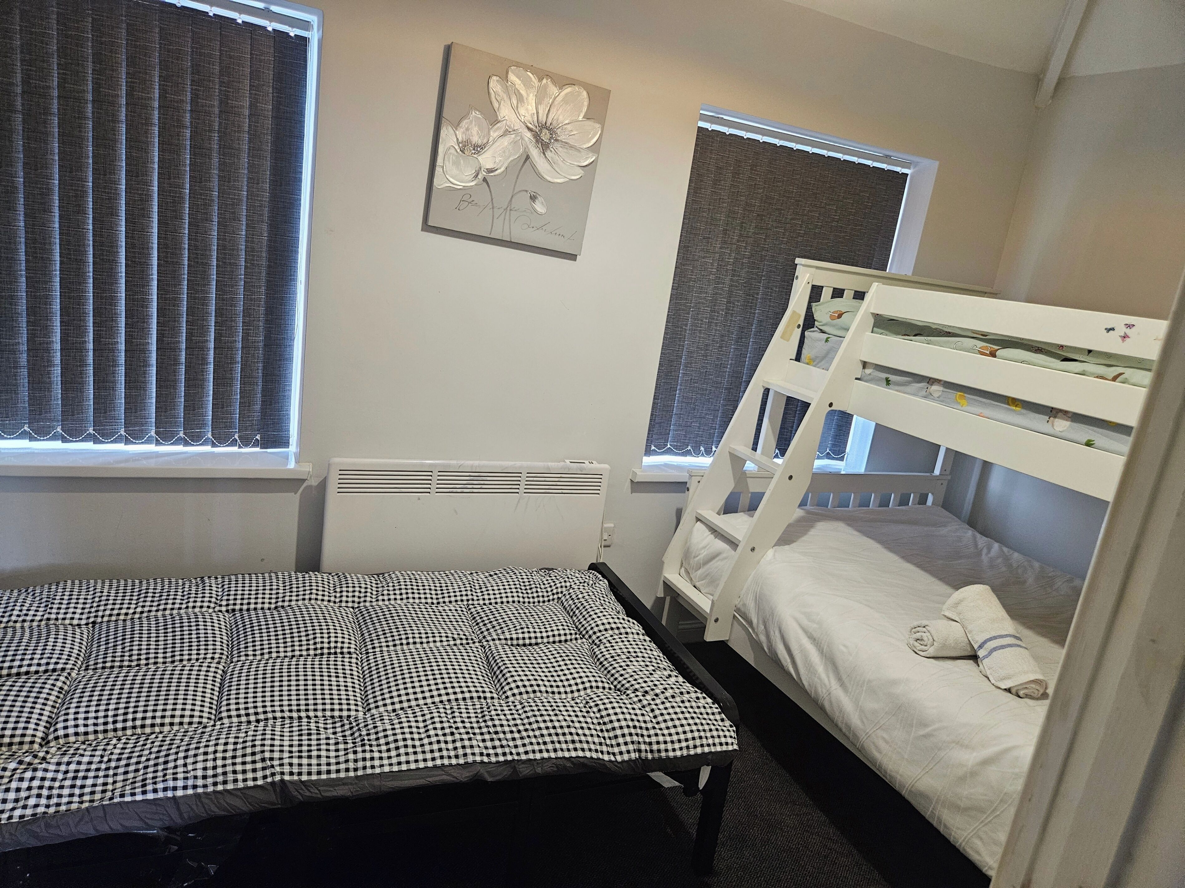 Un dormitorio con una cama litera, una colcha a cuadros, un radiador y una ventana con persianas.