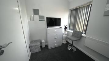 Habitación de hotel compacta con cama, escritorio, silla, televisor, planta en maceta y ventana con persianas.