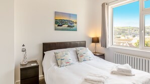 A bedroom with a bed, a nightstand, a lamp, a painting, and a view of a town through a window. - 2 bedroom accommodation in Brixham (Brixham)