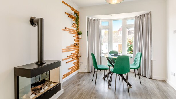 A modern dining area with a glass table, green chairs, a fireplace, and a view of the outside through large windows. - 2 bedroom accommodation in Brixham (Brixham)