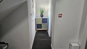 Un couloir étroit avec une petite plante posée sur un meuble, un détecteur d’incendie et une poubelle.