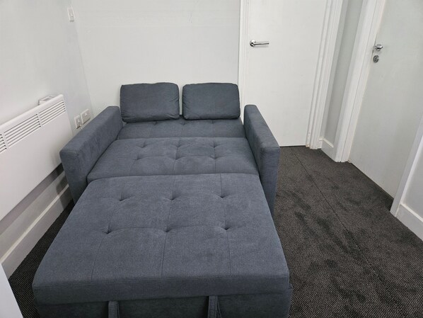 Un sofá cama gris con dos almohadas, un radiador y una puerta en una habitación con piso alfombrado.