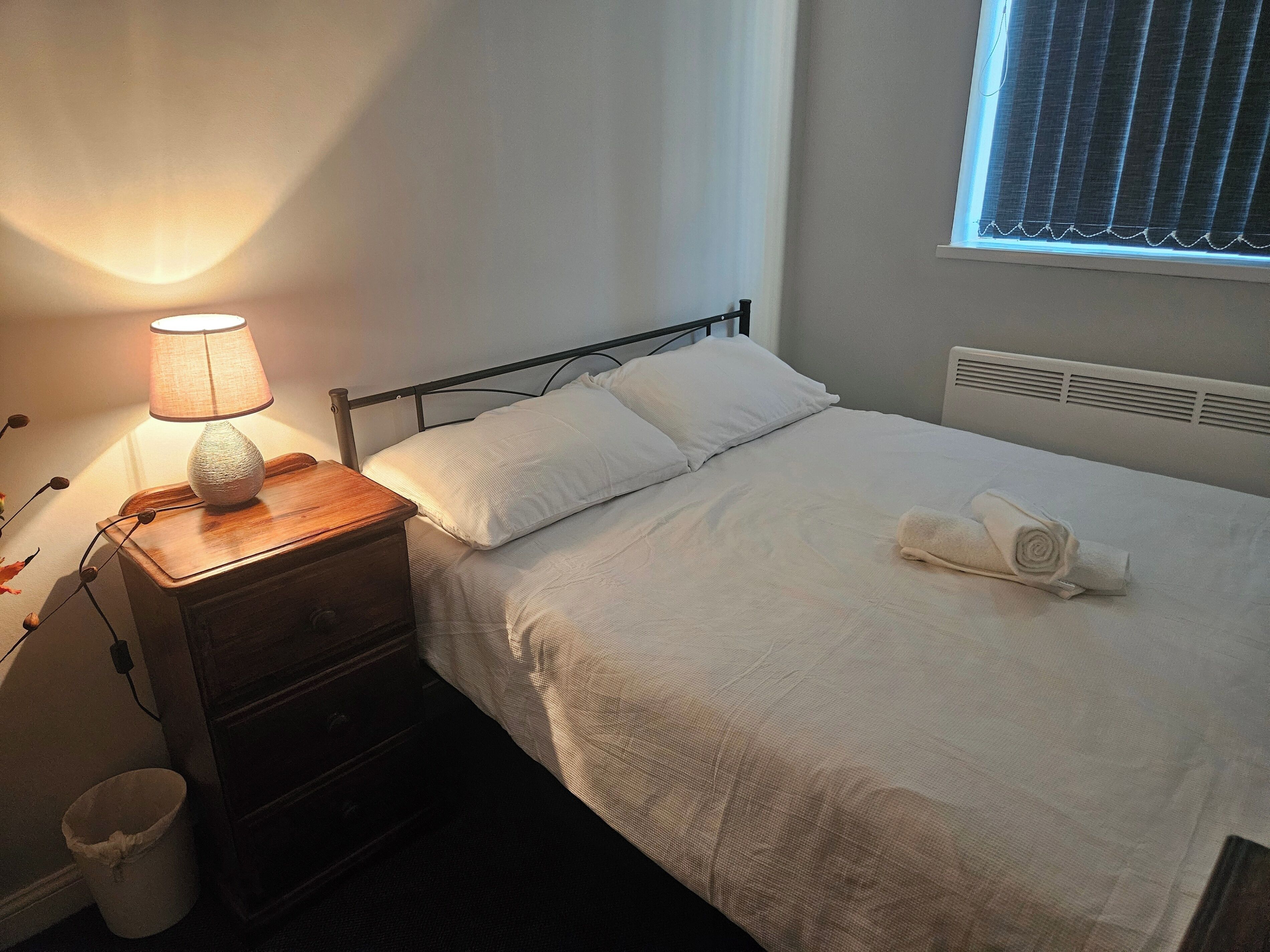 Un lit bien fait, avec du linge de lit blanc, une lampe de chevet et une serviette enroulée posée sur le lit.