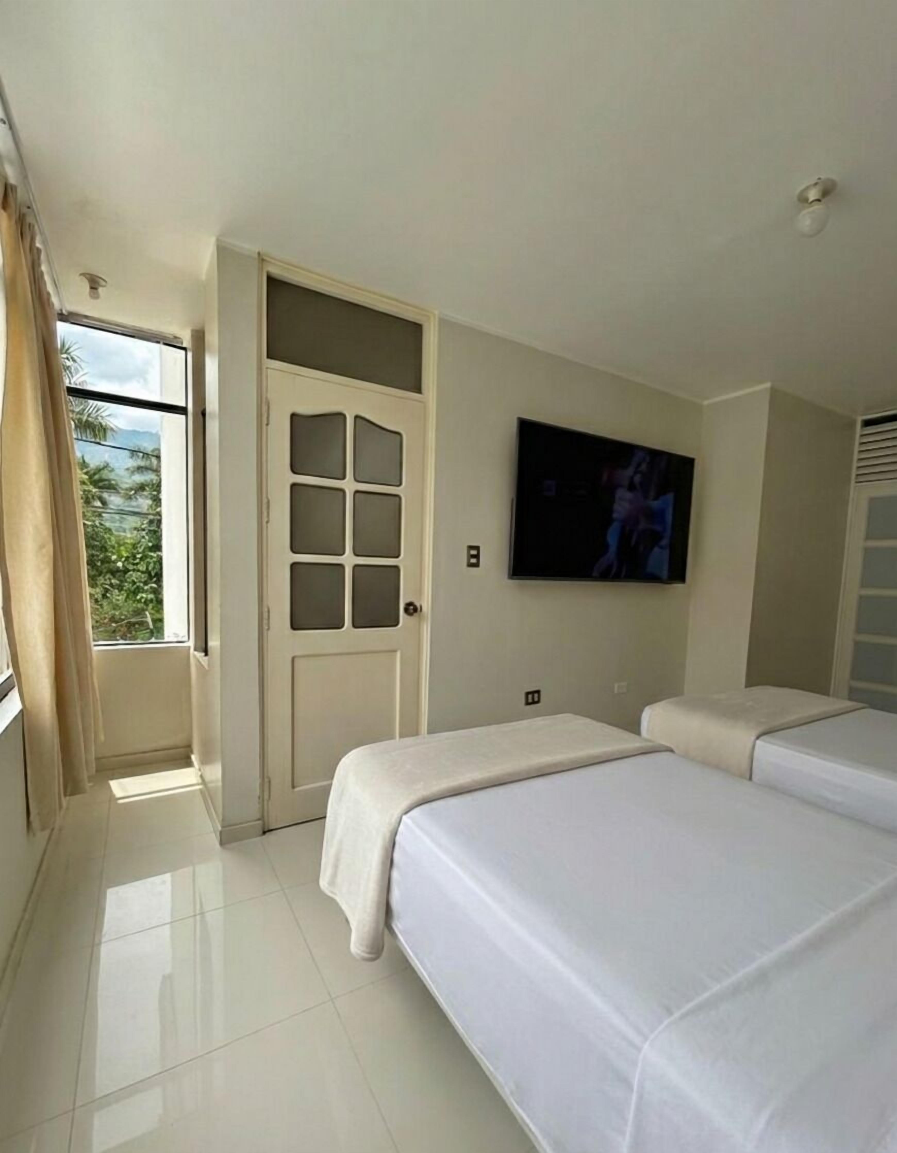 Habitación de hotel con un gran ventanal, una puerta con panel de vidrio satinado, un televisor de pantalla plana montado en la pared y dos camas con ropa blanca.