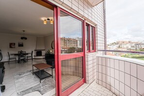 Un balcón con puerta de vidrio de marco rojo, una zona de comedor con mesa y sillas, y vistas de la ciudad.