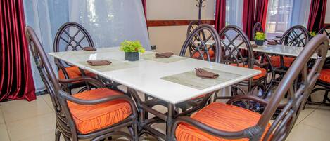 A dining area with a white table surrounded by wicker chairs with orange cushions, a small plant on the table, and red curtains.