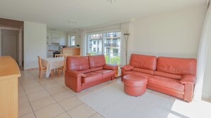 A modern living room with a large red sofa, a dining table with chairs, and a kitchen area visible through an open door. - Apartment Near Belgian Coast With Terrace (Koksijde)