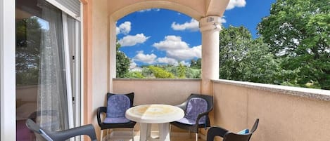 A balcony with a table and four chairs, a sliding glass door, and a view of trees and sky.