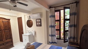 A bedroom with a bed, a ceiling fan, a window with curtains, a wooden wardrobe, and a bathroom with a sink and mirror. - La Casa del Árbol - Oaxaca Historic Center (Oaxaca de Juárez)