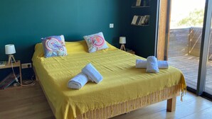 A bed with a yellow bedspread and two decorative pillows, a bedside table with a lamp, and a view of the outdoors through a glass door. - Charming house in the greenery - Downtown Porto-Vecchio (Porto-Vecchio)