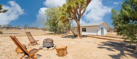 A desert landscape with a white house, two wooden lounge chairs, a fire pit, and a tree.