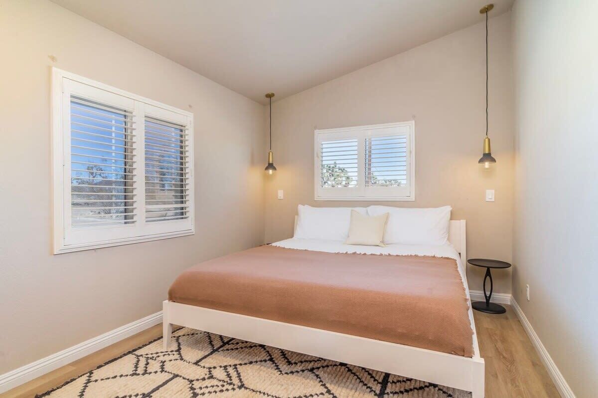 Un dormitorio con cama, dos ventanas con persianas, una mesita de noche y una alfombra.