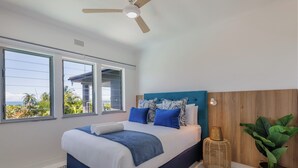 Une chambre avec un lit, de grandes fenêtres, un ventilateur de plafond et une vue sur des palmiers.