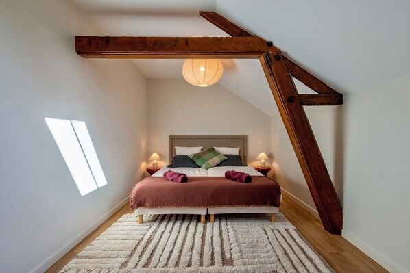 A bedroom with a sloped ceiling, a large bed with two pillows, a hanging light fixture, and a window providing natural light. - Domaine de Fontenay (Fontenay-sur-Vègre)