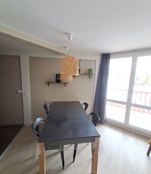 A dining area with a table and chairs, a hanging light fixture, a plant on a shelf, and a view through a large window. - T2 apartment in residence with pool (Le Touquet-Paris-Plage)