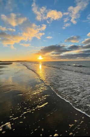 Une scène de plage au coucher du soleil, avec le soleil qui disparaît à l’horizon, des vagues qui viennent doucement lécher le rivage et un ciel dégagé parsemé de quelques nuages.