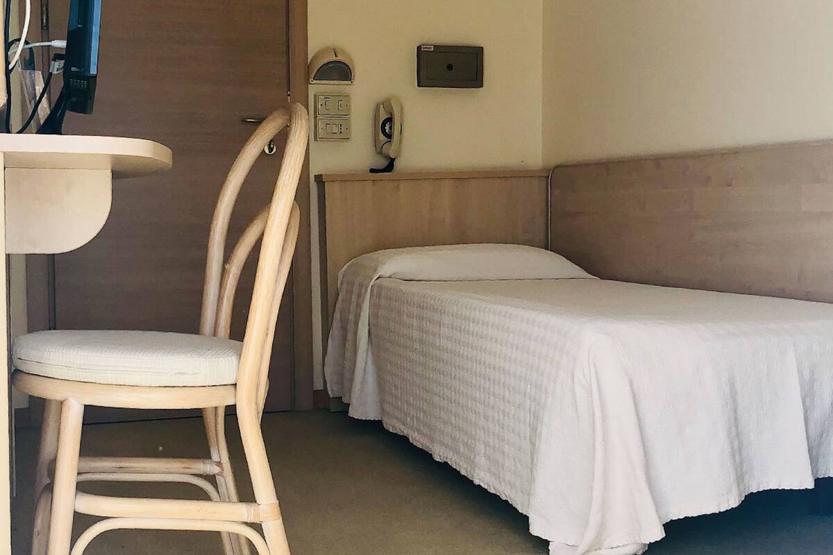 Una cama individual con cabecera de madera, una silla con cojín, una mesita y un teléfono instalado en la pared.