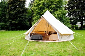 A tent with a bed and a small table inside, surrounded by grass and trees. - Nine Yards Bell Tents - Coleraine (Coleraine)