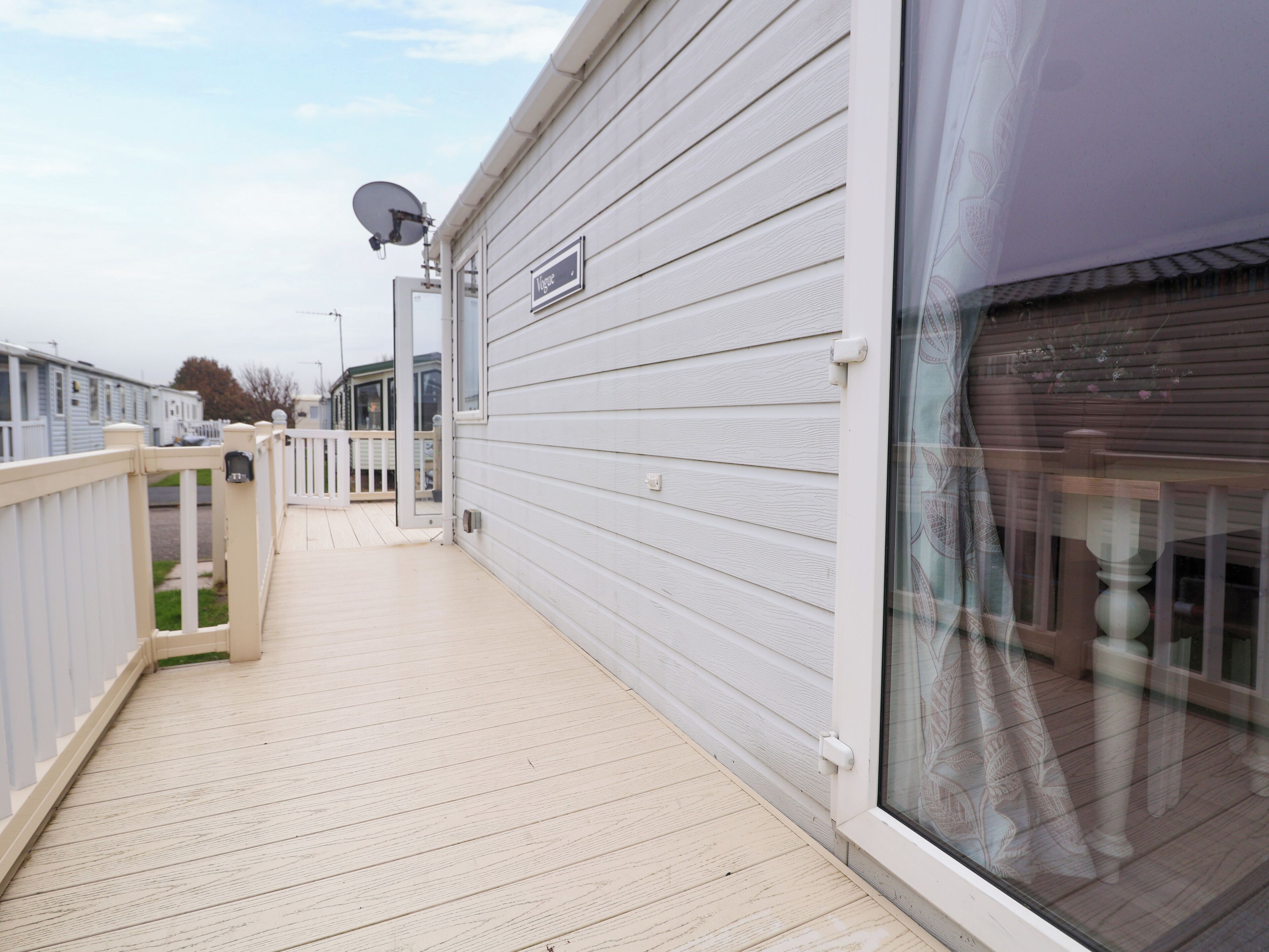 Une terrasse en bois avec une balustrade blanche, une antenne parabolique et une fenêtre agrémentée d’un rideau décoratif.