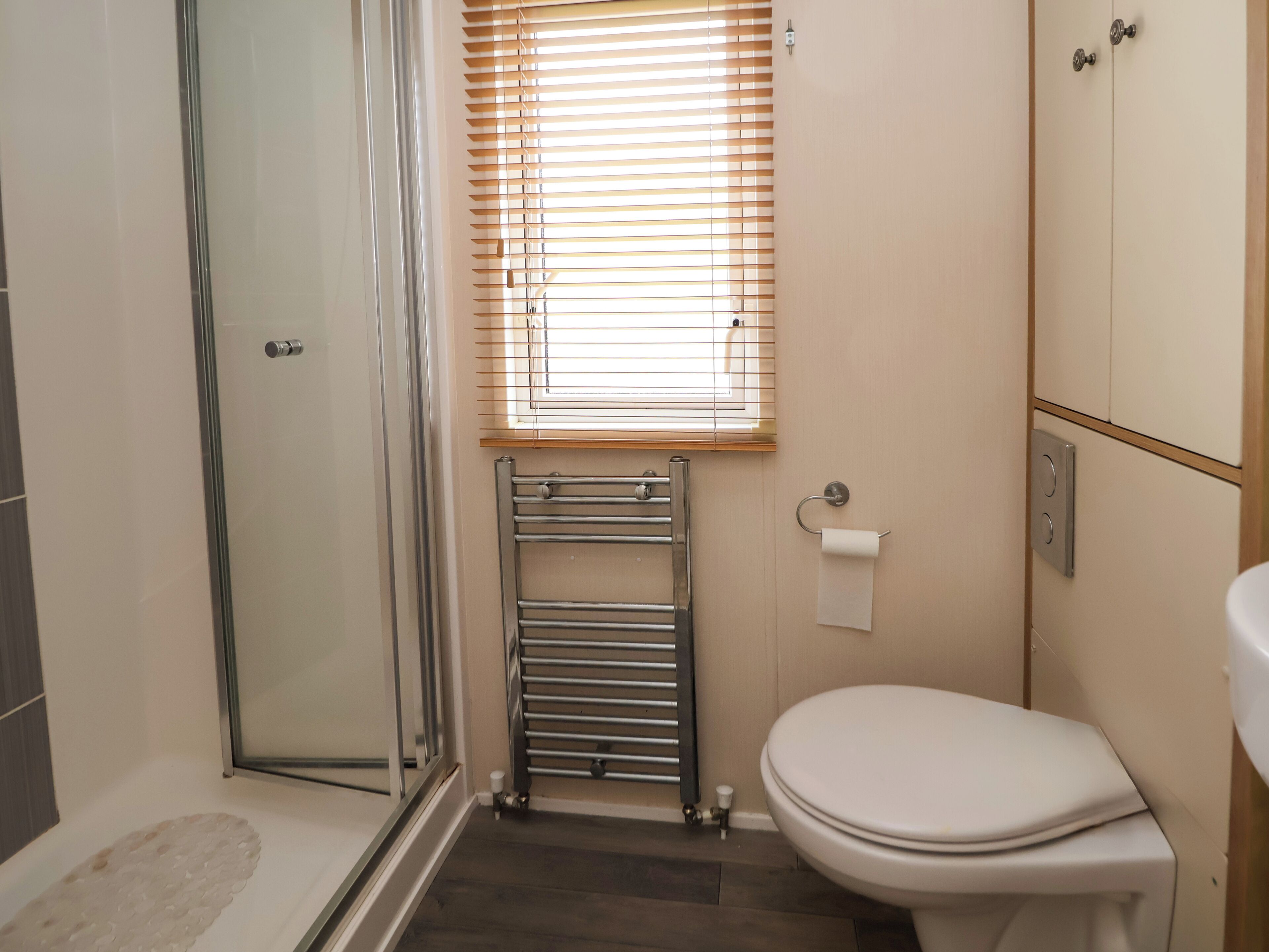 A bathroom with a glass shower enclosure, a toilet, a towel rack, and a window with blinds.