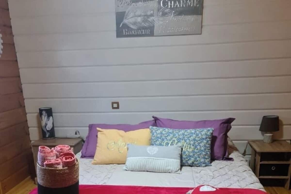 A neatly made bed with a red blanket and multiple pillows, a small bedside table with a lamp, and a framed picture on the wall.