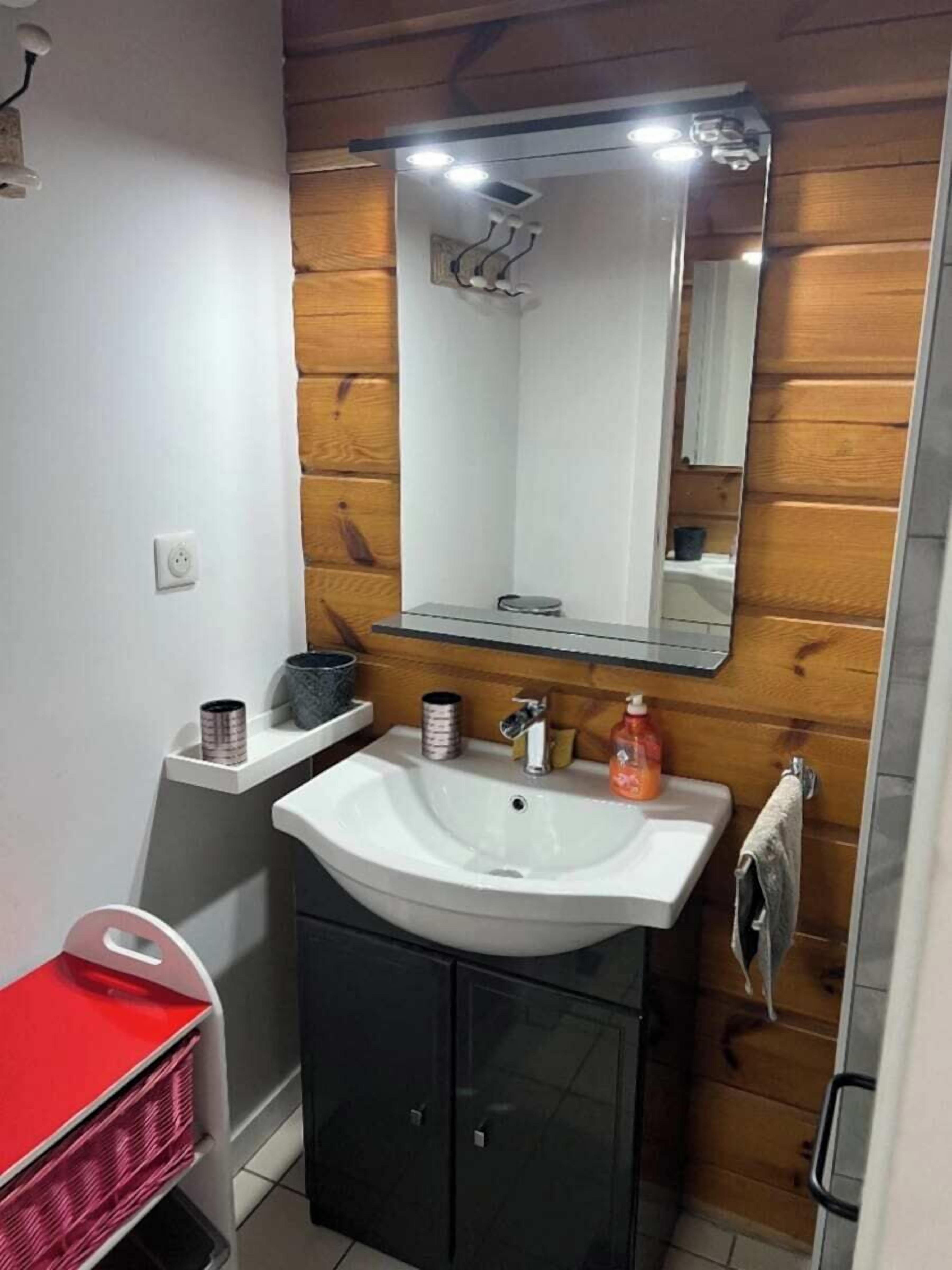 Une salle de bain avec un lavabo blanc, des murs recouverts de panneaux en bois, un miroir au-dessus du lavabo et une chaise rouge.
