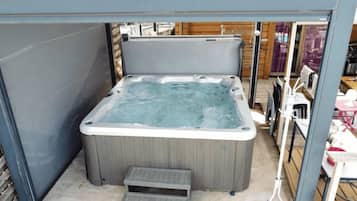 A hot tub with a wooden exterior, a small step leading up to it, and a view of the surrounding area including a wooden deck and outdoor furniture.