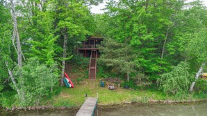 A cabin with a wooden staircase leading up to a deck, surrounded by dense forest. - Make Good Choices! LAKE FRONT- 2 King beds and 2 baths (waupaca)